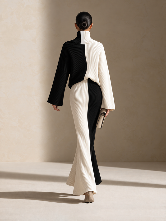 A model is shown from behind wearing a high-neck, long-sleeve ribbed knit two-piece set in black and off-white, paired with beige heels and holding a small clutch book.