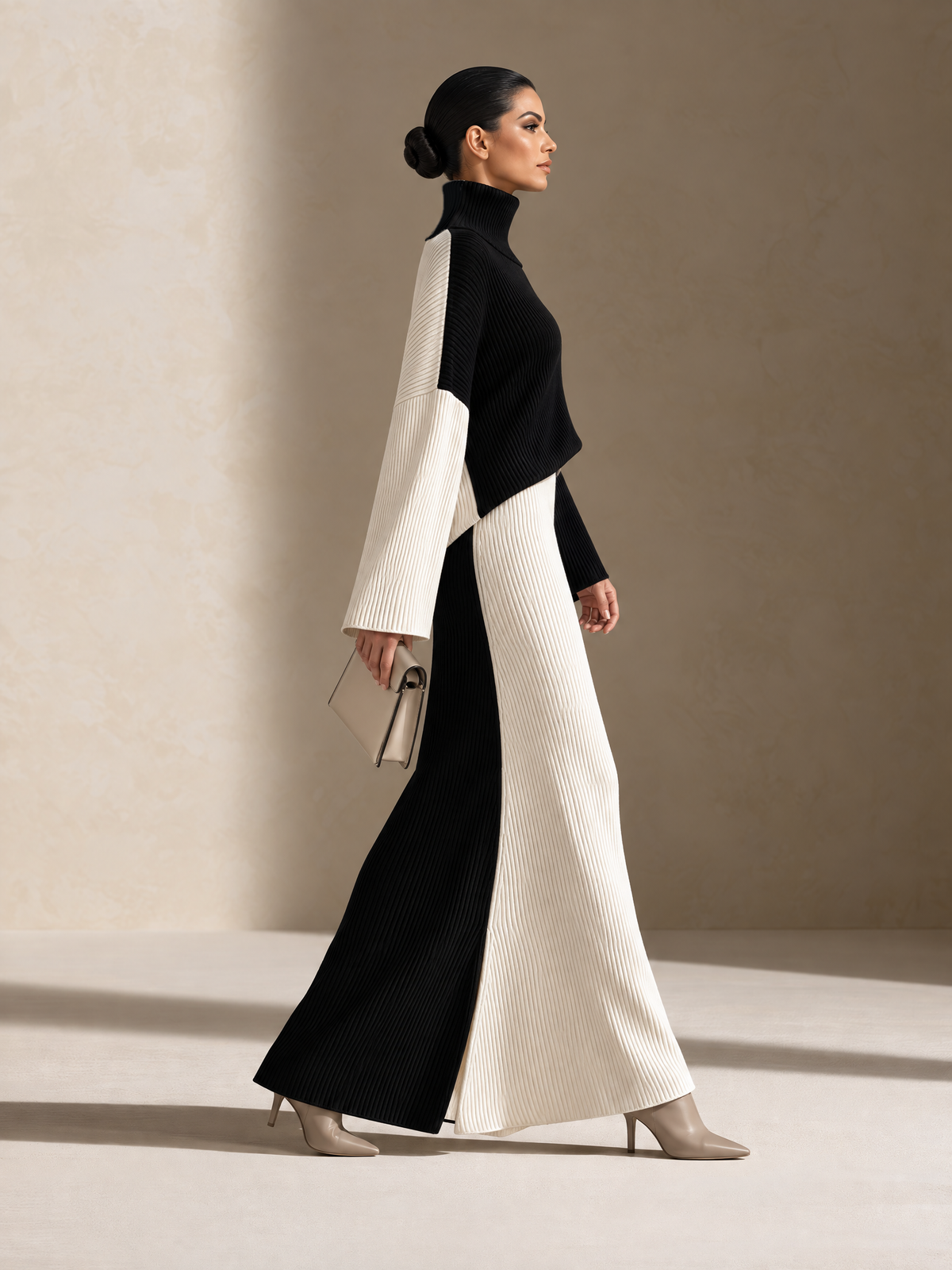 A woman models a long-sleeved, high-neck maxi dress with a bold black and off-white color-blocked design. The dress features a ribbed texture on the lower skirt and a smooth matte finish on the sleeves and bodice.