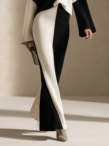 A model wearing a long, pleated color-block skirt in black and cream, paired with a matching cream and black top, holding a small beige clutch against a warm beige background.